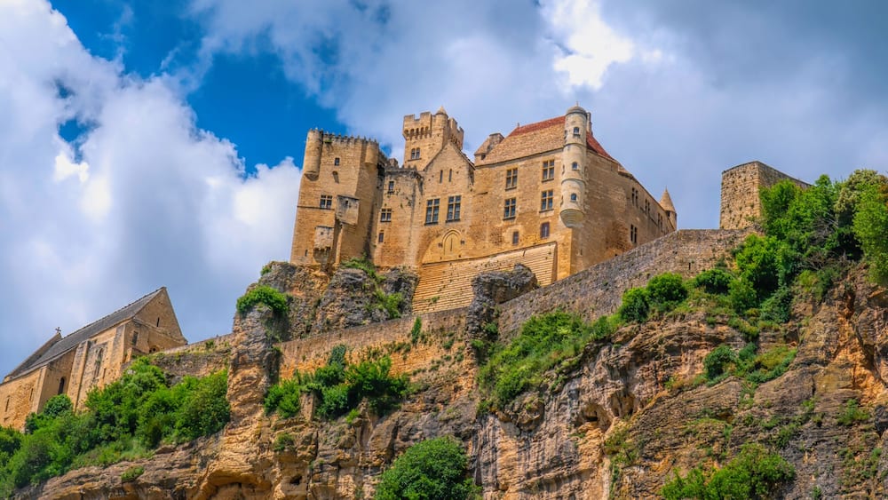 Un chĂąteau historique perchĂ© au sommet dâune colline rocheuse, entourĂ© dâune vĂ©gĂ©tation luxuriante, sous un ciel partiellement nuageux.