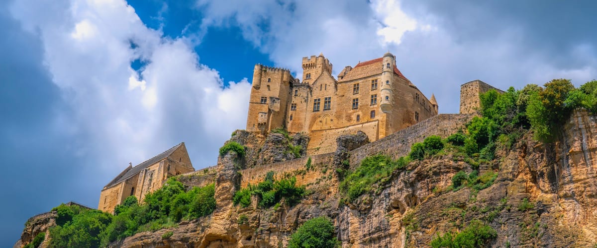 A historic castle perched atop a rocky hill with lush greenery, under a partly cloudy sky.