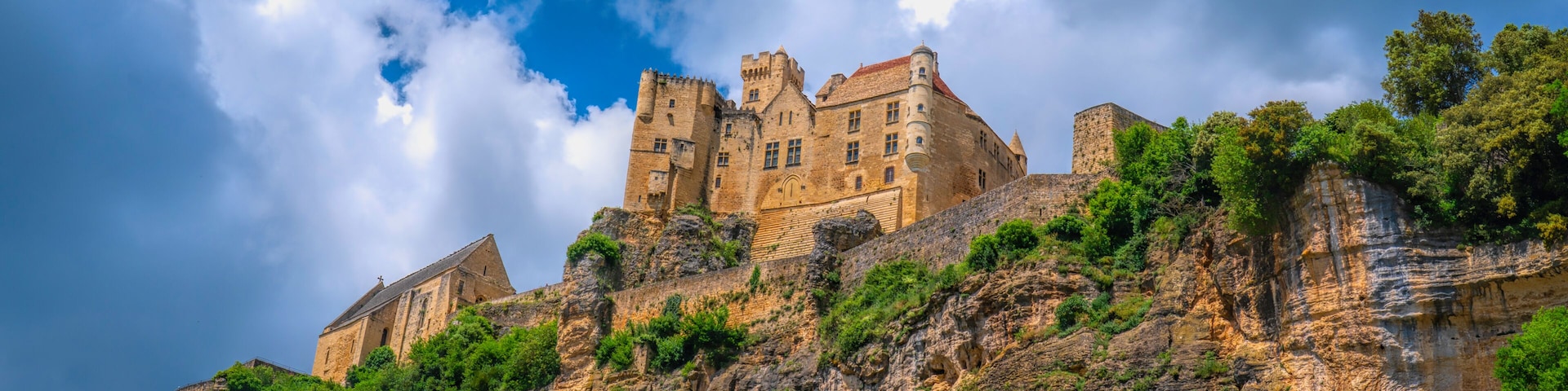 A historic castle perched atop a rocky hill with lush greenery, under a partly cloudy sky.