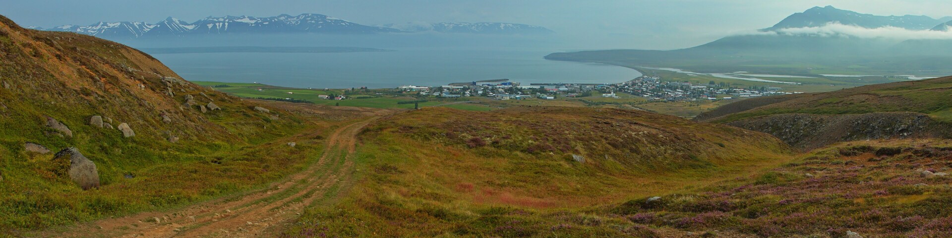 A coastal town with a dirt path leading to it, surrounded by hills and mountains.