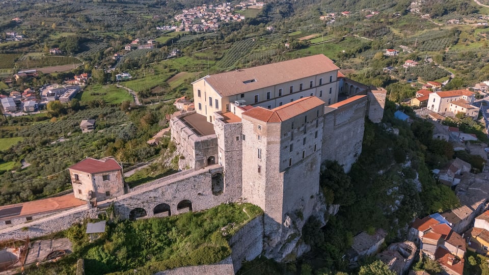 A historic stone castle with a red-tiled roof, surrounded by lush greenery and a rural village in the background.