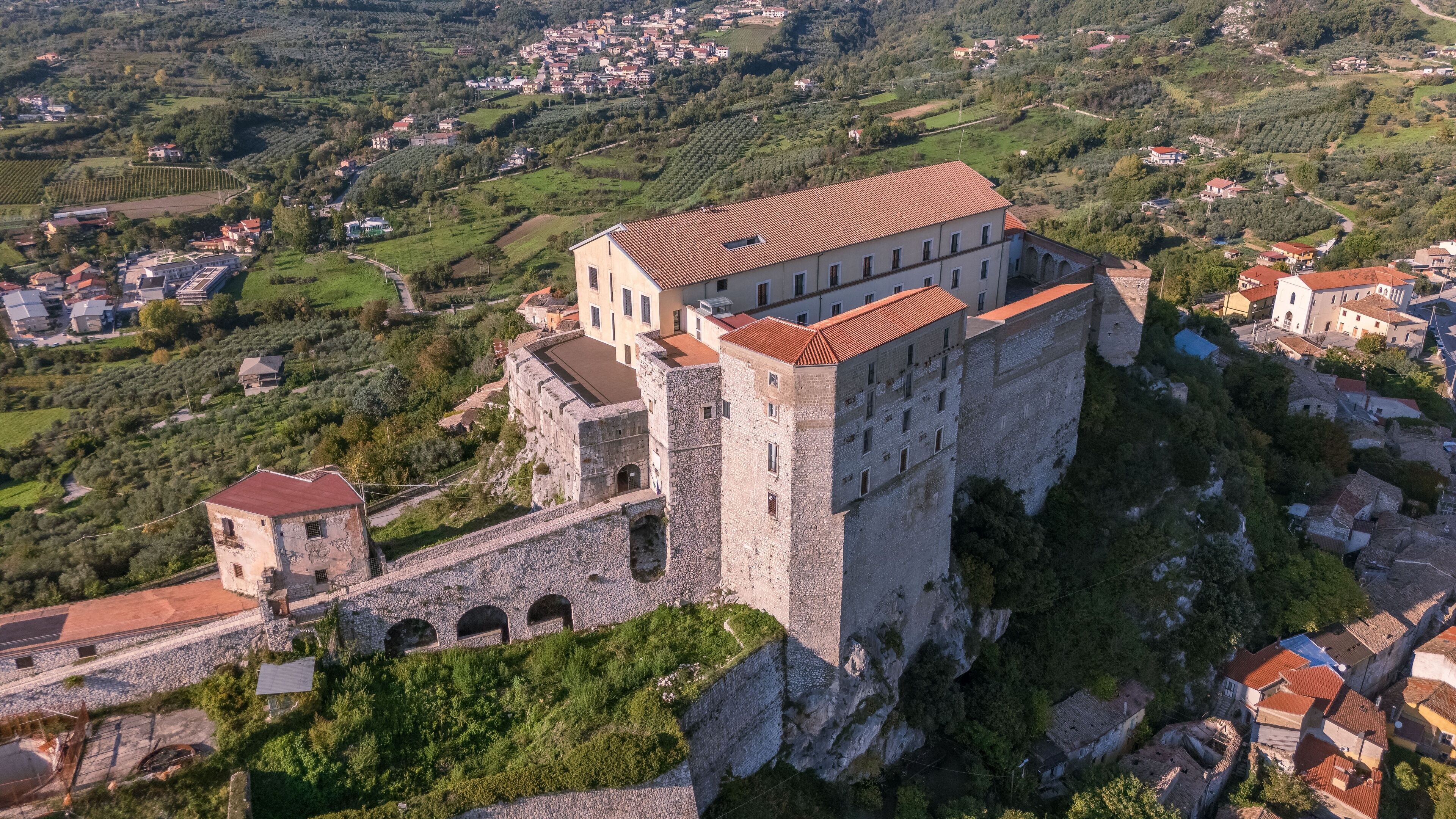 A historic stone castle with a red-tiled roof, surrounded by lush greenery and a rural village in the background.