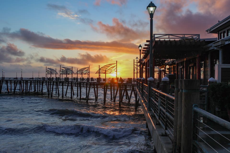 A pier extending into the ocean at sunset with a street lamp and buildings on the right.
