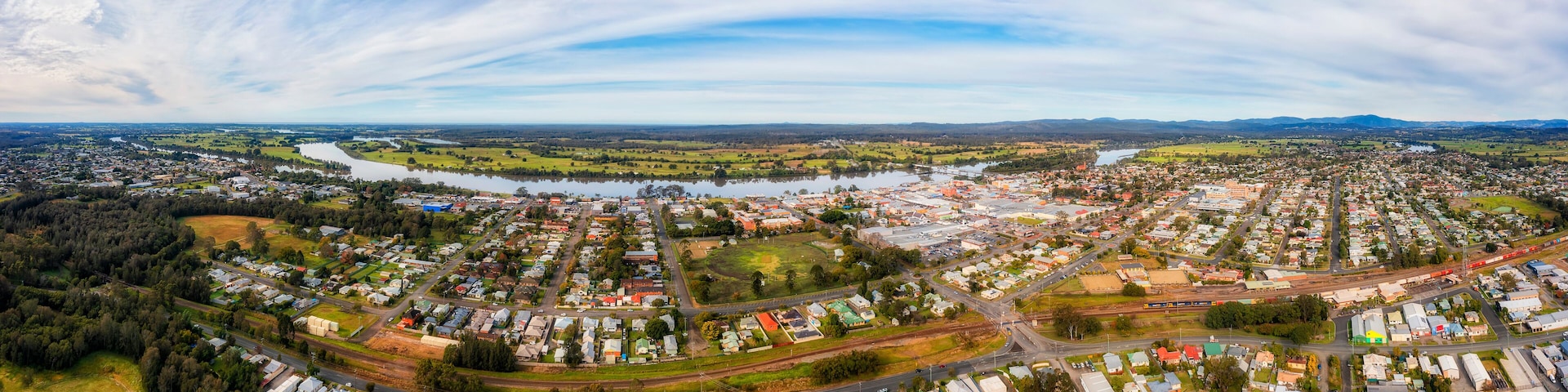 A panoramic view of a town with a river, green fields, and a mountainous backdrop.