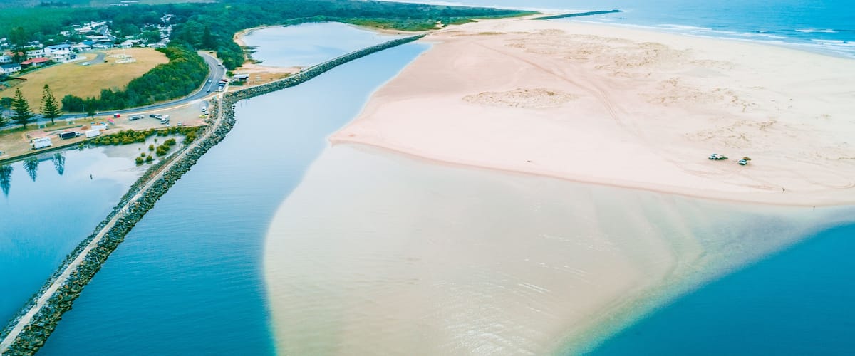 A coastal landscape with a sandy beach, a calm river, and a small settlement with buildings and greenery.