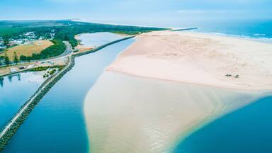 A coastal landscape with a sandy beach, a calm river, and a small settlement with buildings and greenery.