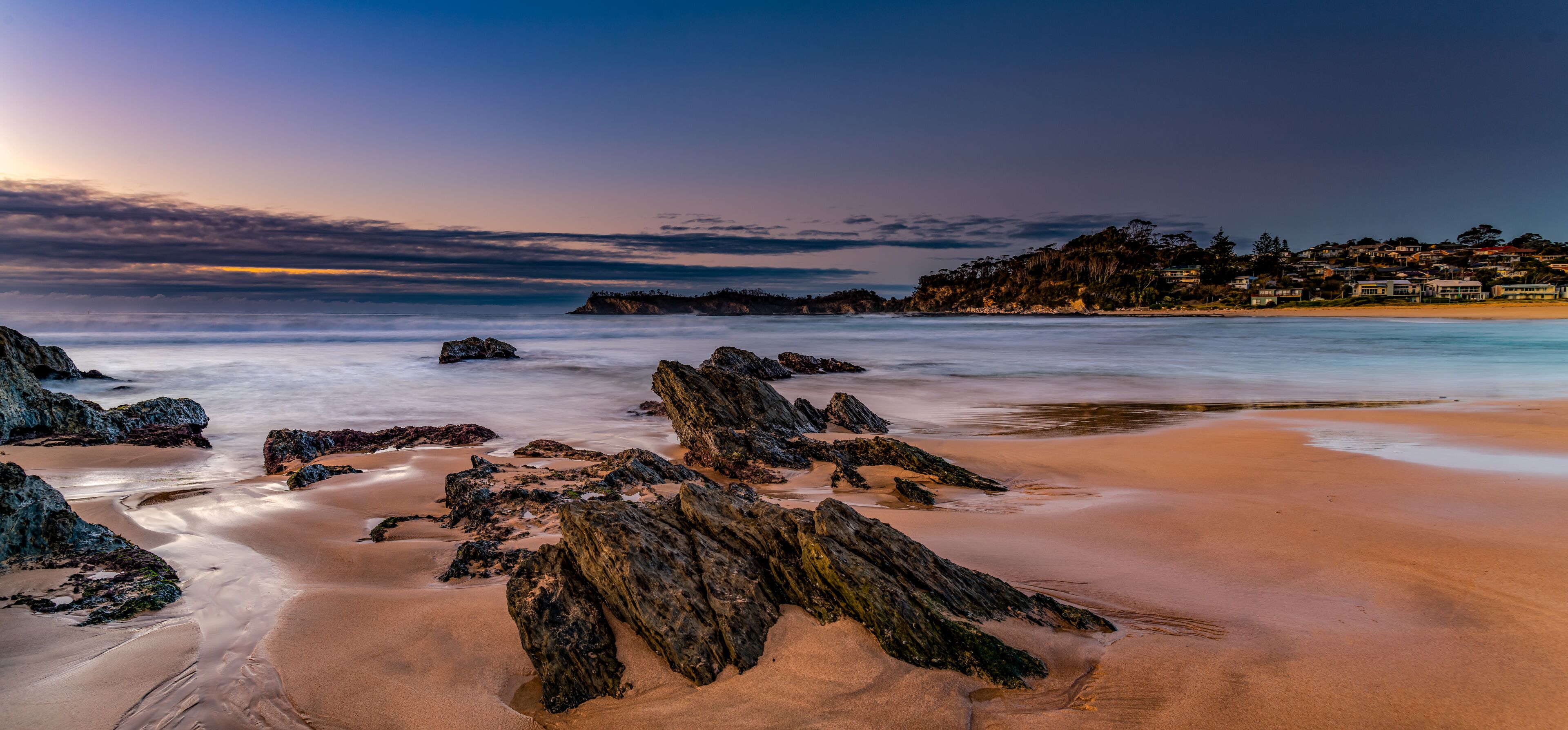 Paisagem costeira com praia arenosa, rochas imponentes e uma linha de costa distante com edifícios.