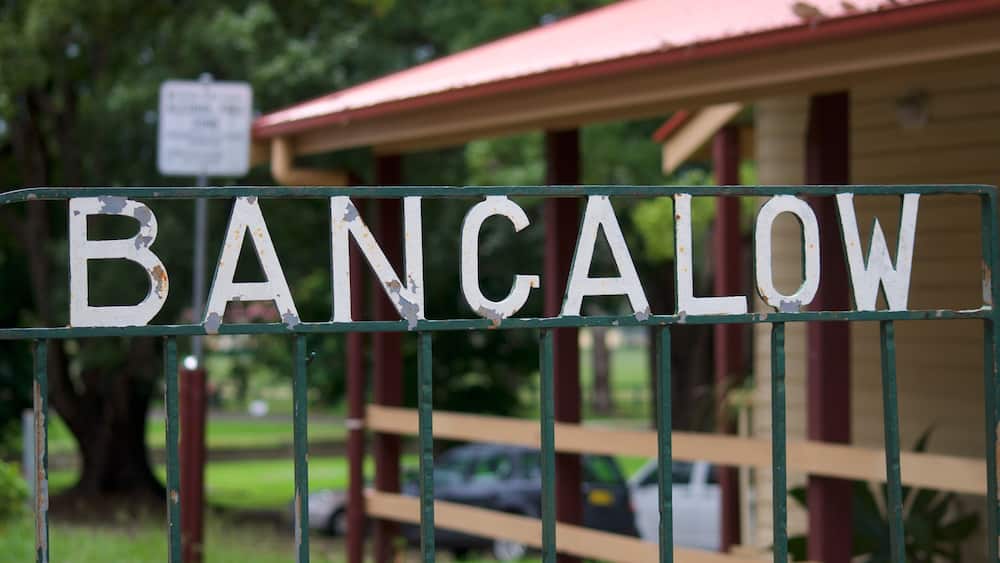 A weathered sign with the word 'BANCALOW' on a metal fence, a red-roofed building, and a car in the background.