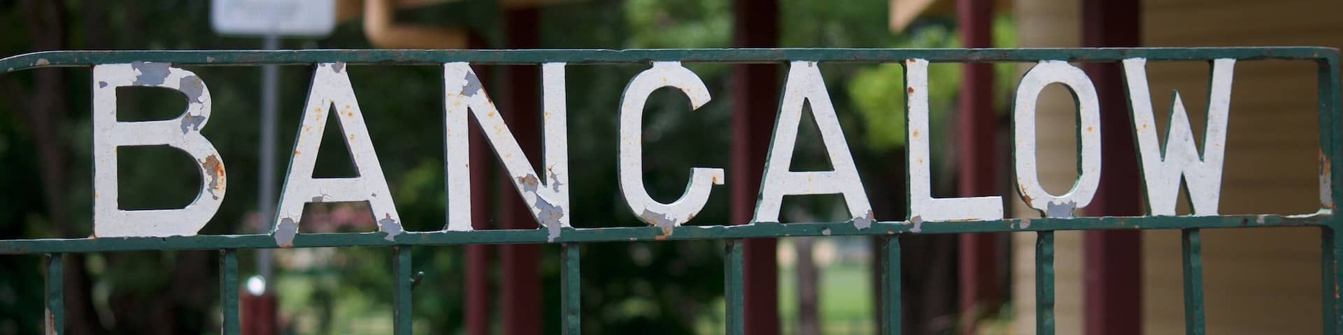 A weathered sign with the word 'BANCALOW' on a metal fence, a red-roofed building, and a car in the background.