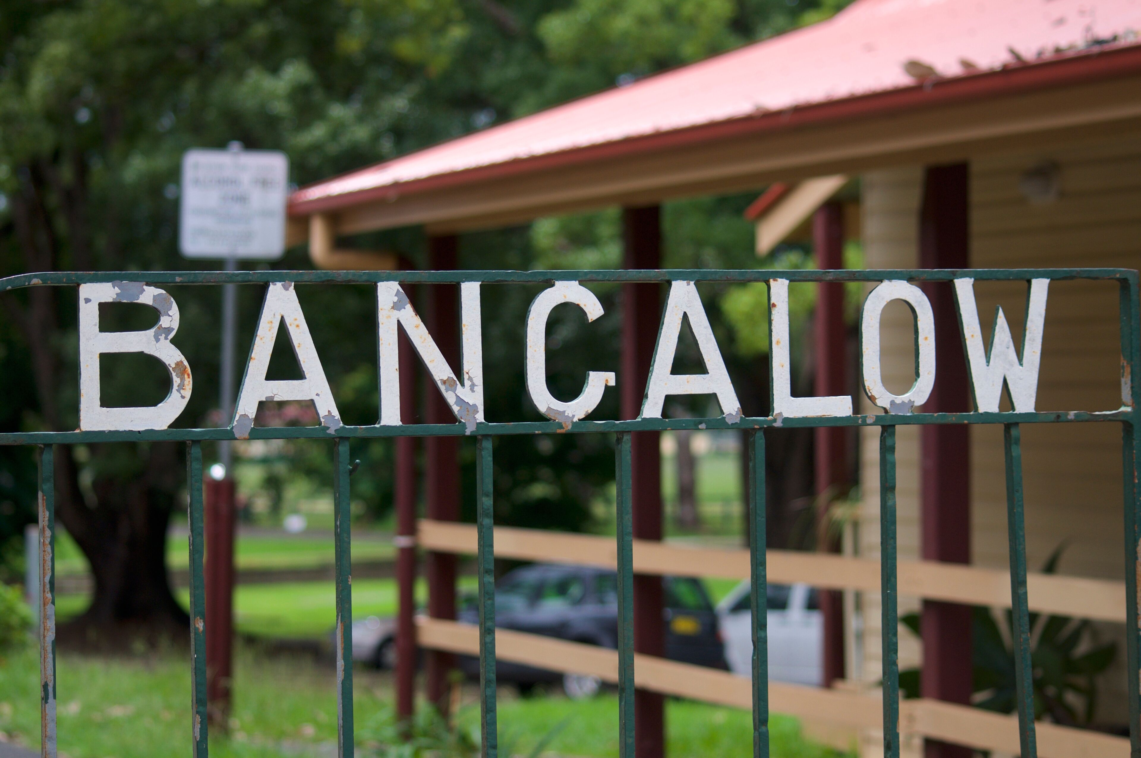 A weathered sign with the word 'BANCALOW' on a metal fence, a red-roofed building, and a car in the background.
