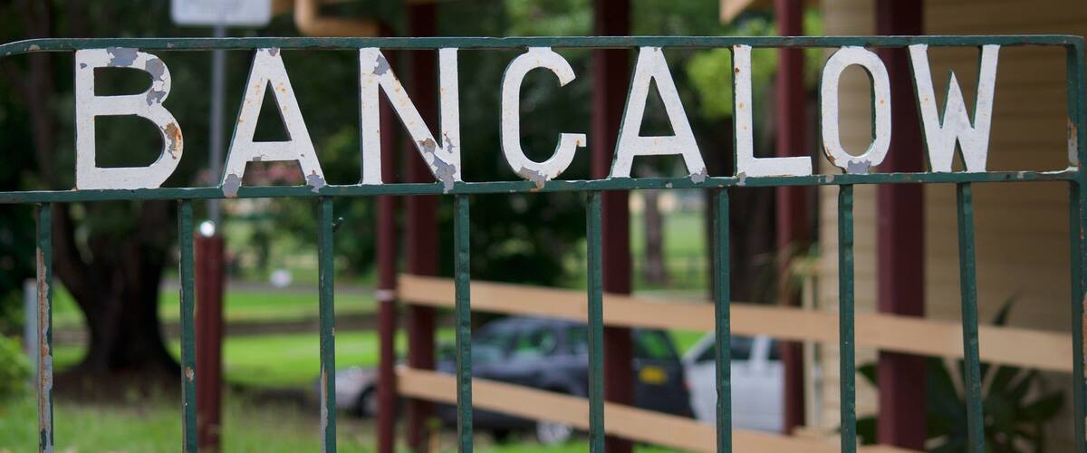 A weathered sign with the word 'BANCALOW' on a metal fence, a red-roofed building, and a car in the background.