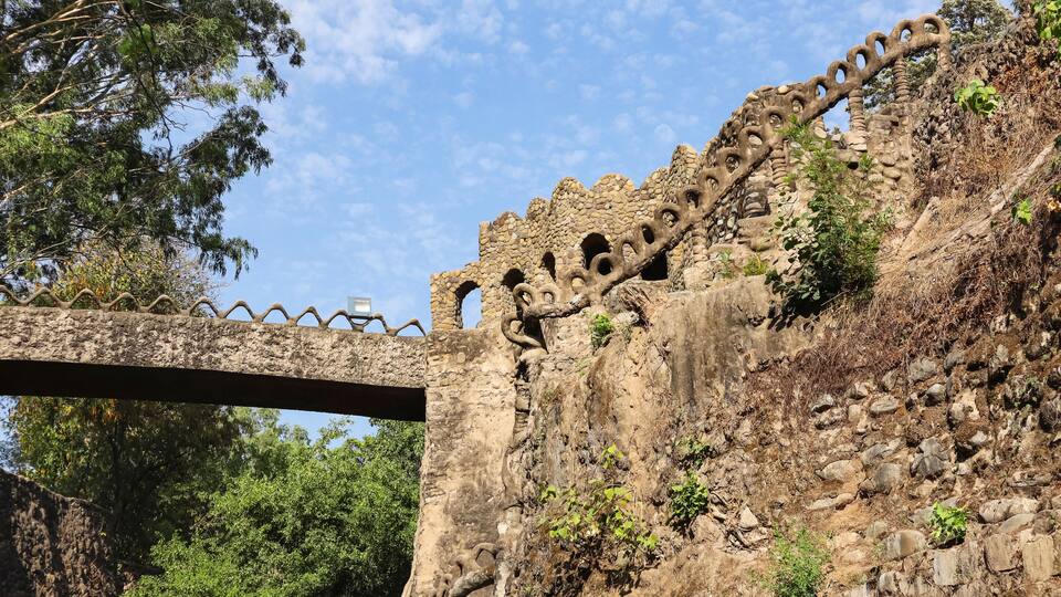 A stone bridge with a unique serpentine design, a stone wall with circular cutouts, and lush green trees in the background.