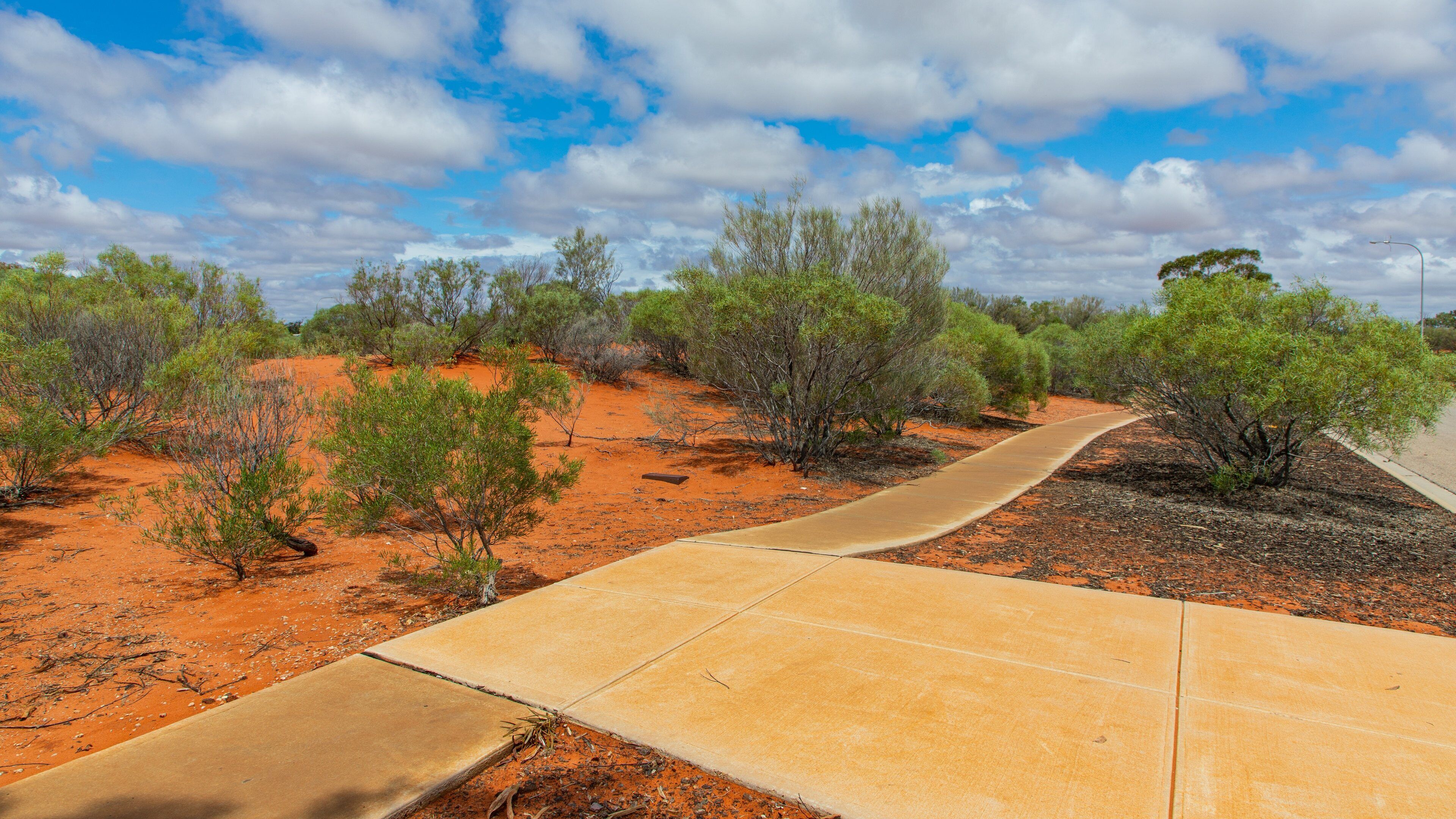 A winding concrete path through a red desert landscape with sparse green vegetation and a partly cloudy sky.