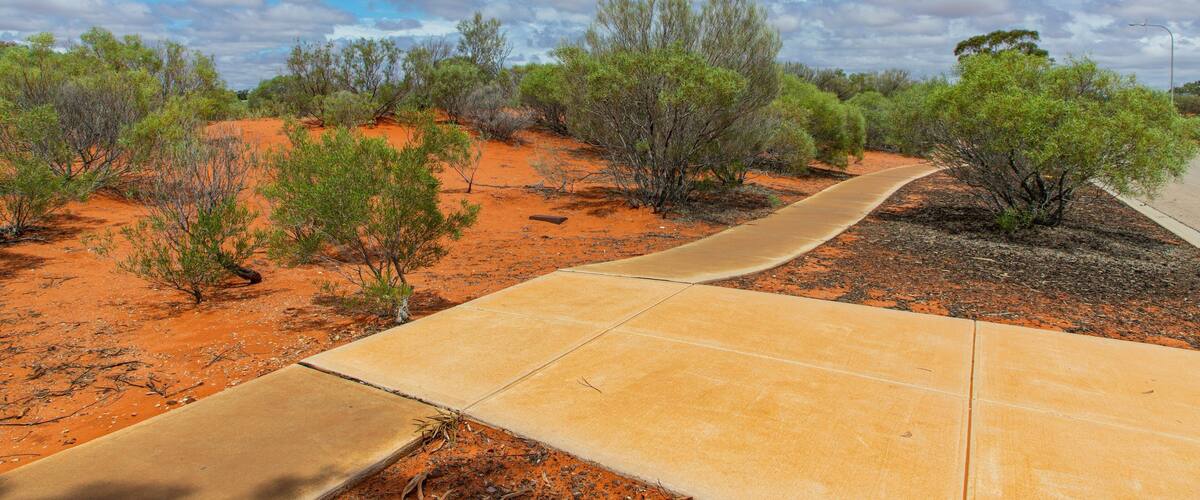 Ein gewundener Betonweg schlängelt sich durch eine rote Wüstenlandschaft mit vereinzelter grüner Vegetation und einem teilweise bewölkten Himmel.