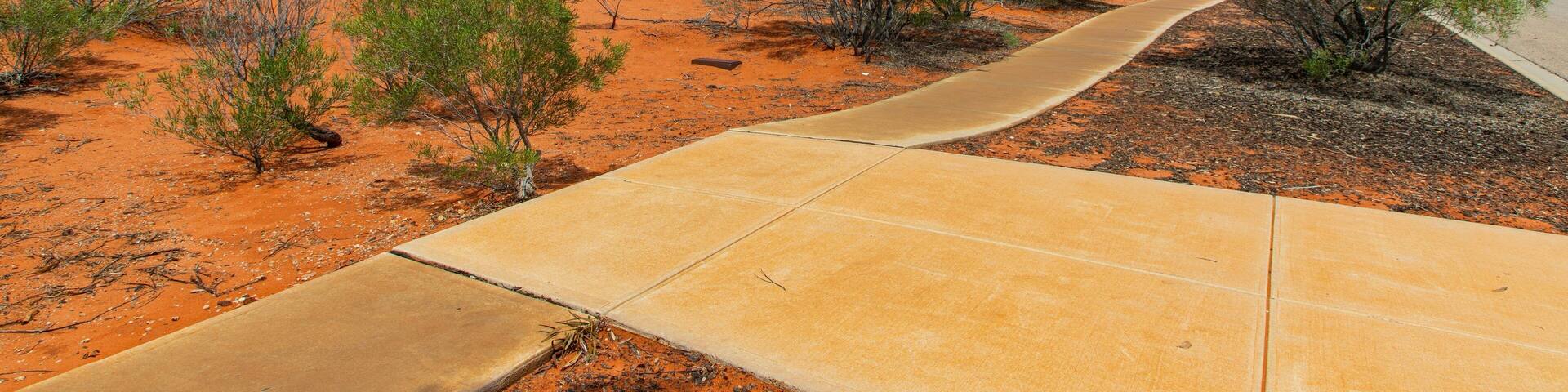 A winding concrete path through a red desert landscape with sparse green vegetation and a partly cloudy sky.
