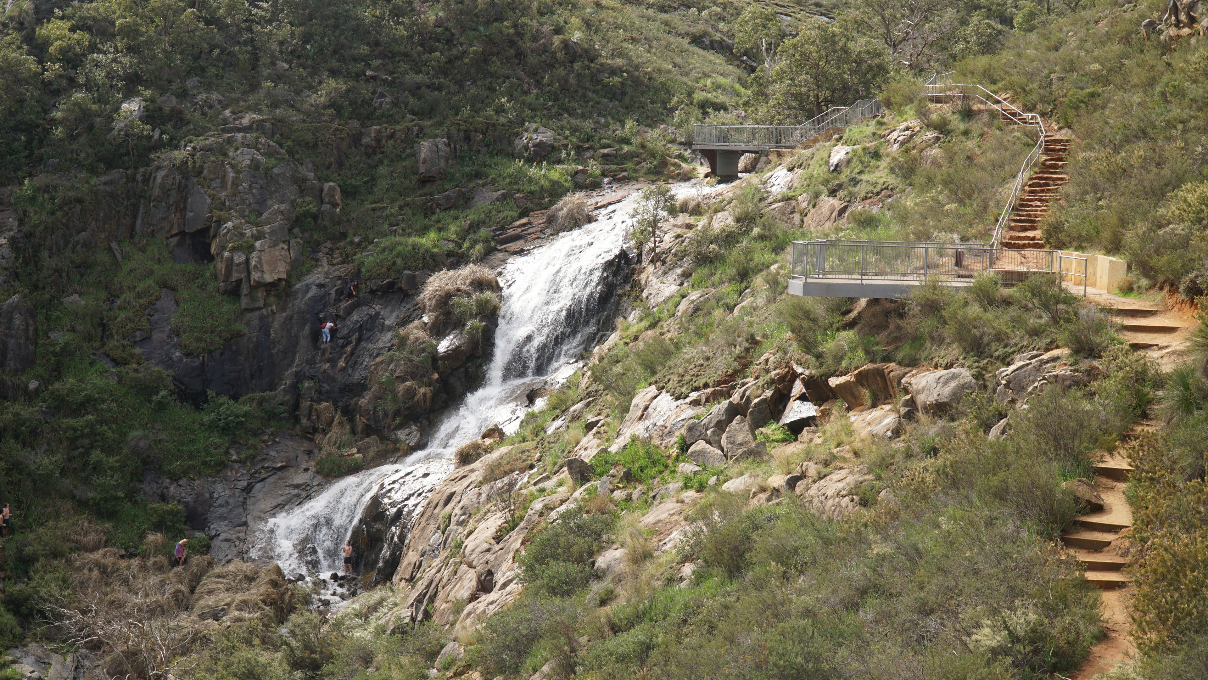 A waterfall cascading down a rocky hillside, a wooden staircase, and a bridge with railings.