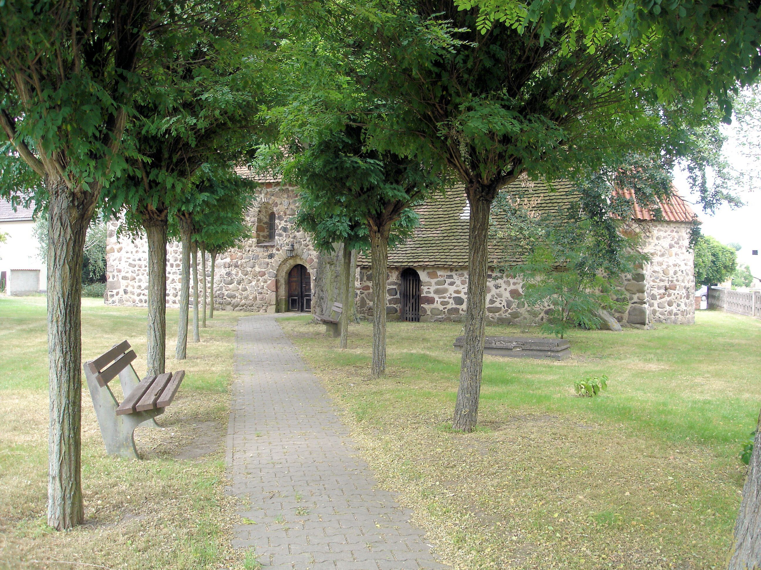 A stone building with a tiled roof, surrounded by trees and a paved pathway.
