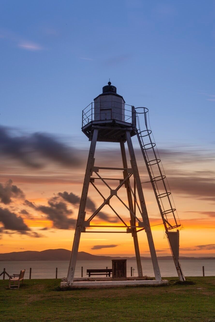 A lighthouse with a metal staircase, a bench, and a grassy area.