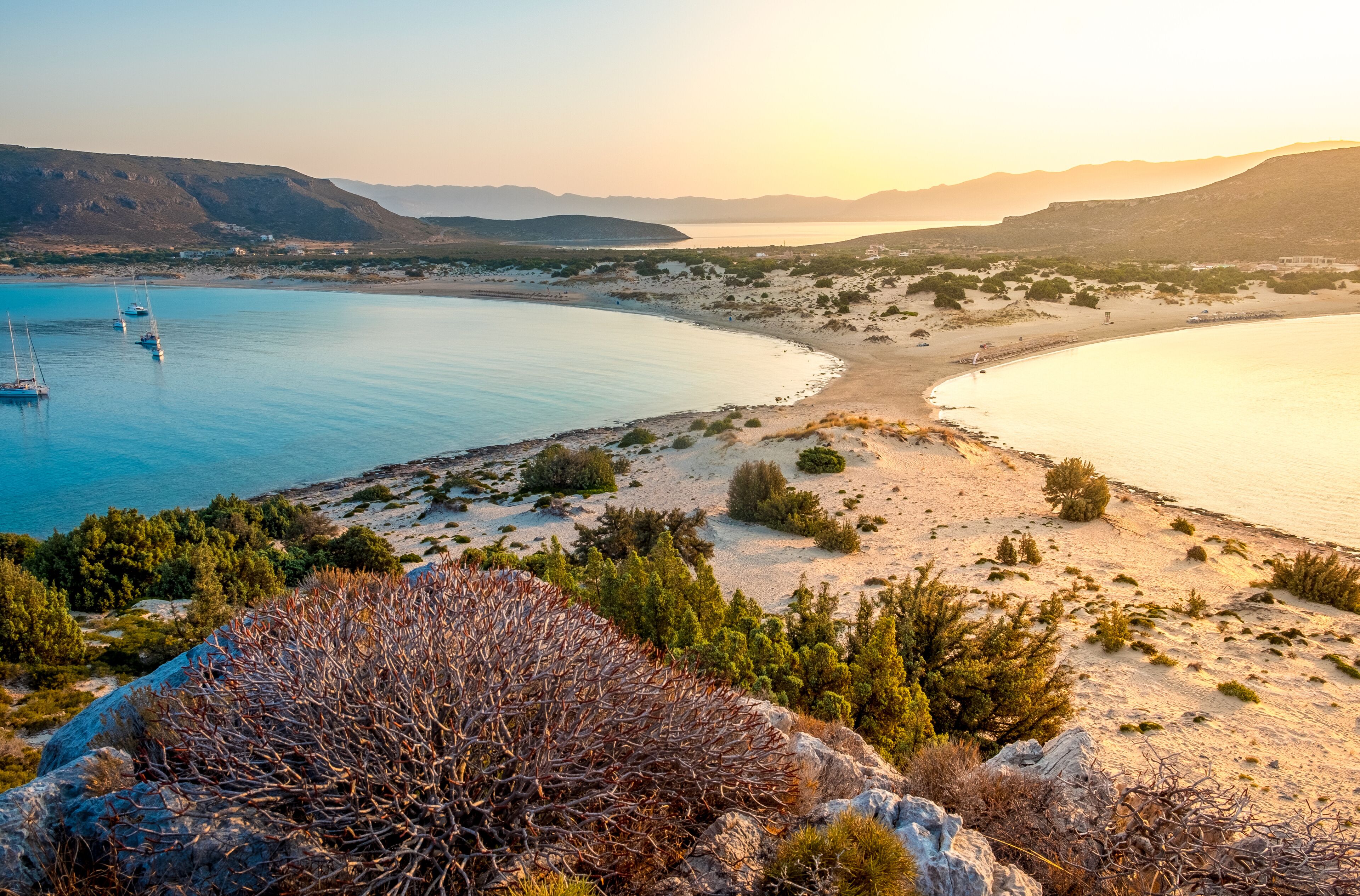 A coastal landscape with a sandy beach, sparse vegetation, and a calm sea with boats.