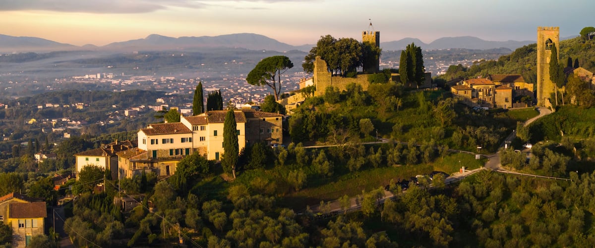 A hillside town with historic stone buildings, a prominent tower, and a view of the distant cityscape.