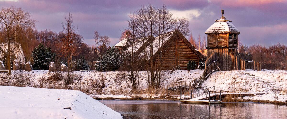 A wooden cabin with a snow-covered roof, a pond, and bare trees in a snowy landscape.