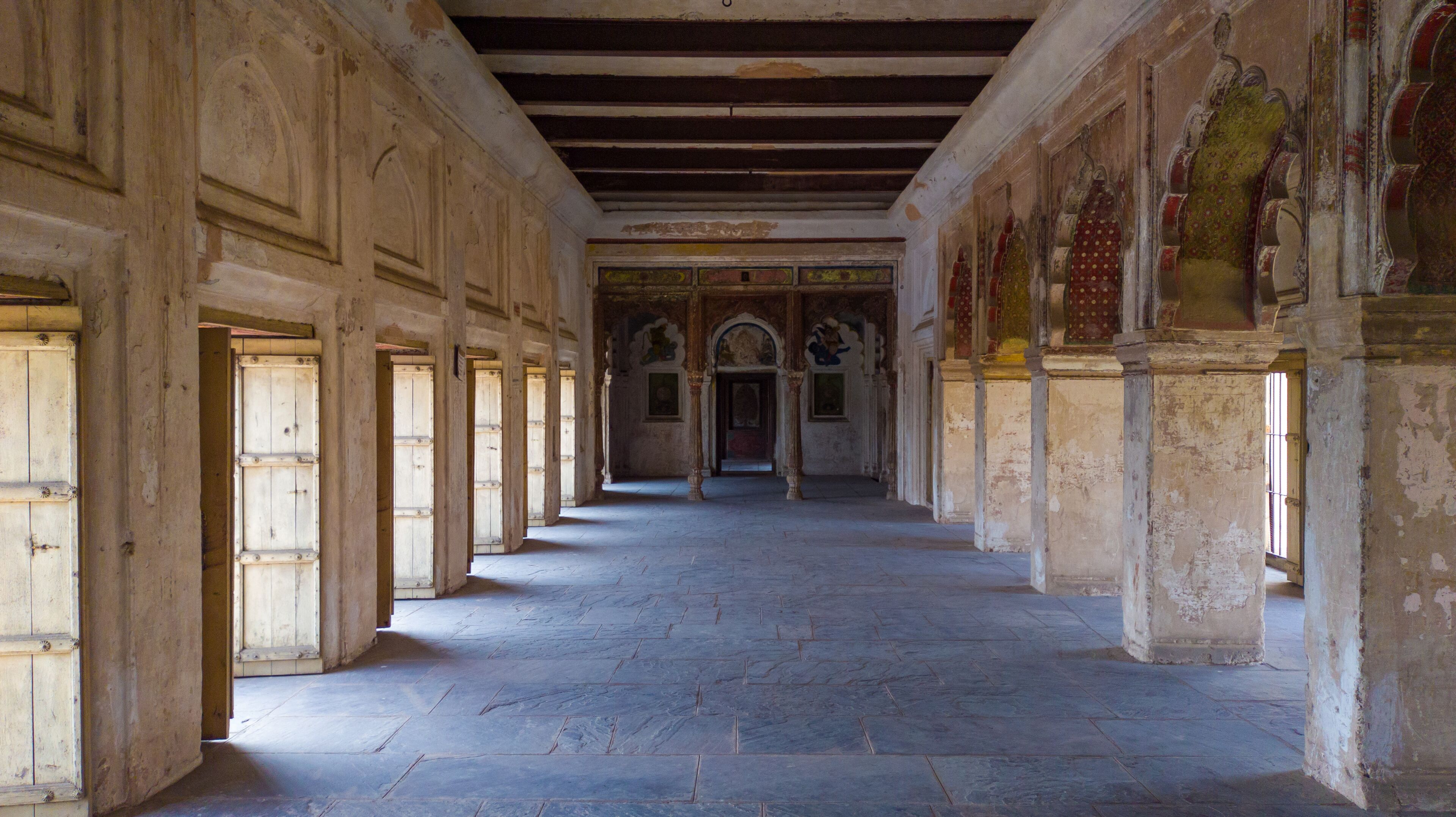 A corridor with stone floors and wooden doors, leading to a central archway.
