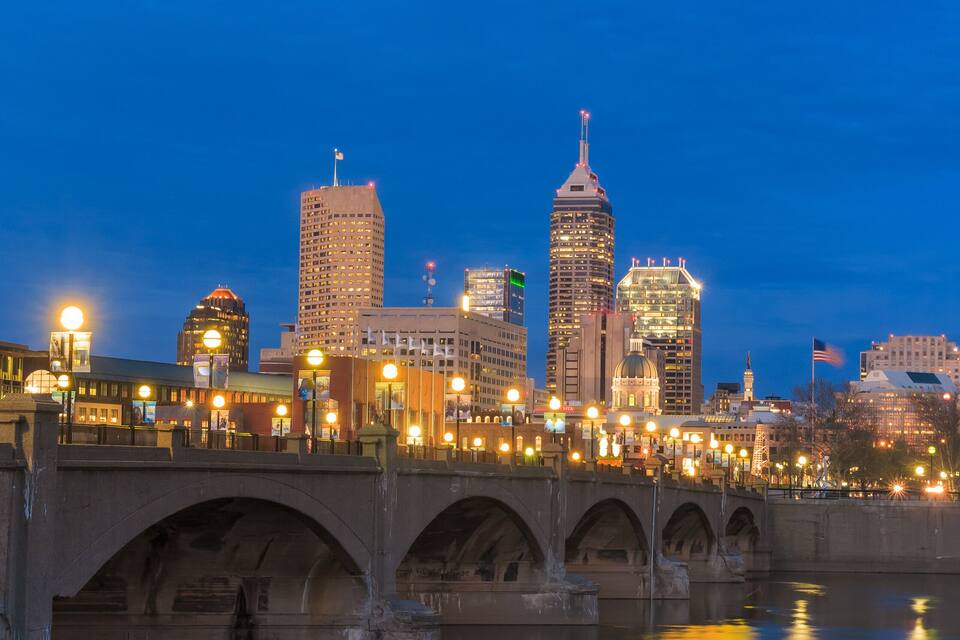 A city skyline at dusk with illuminated buildings, a bridge over a calm river, and a reflection of lights on the water.