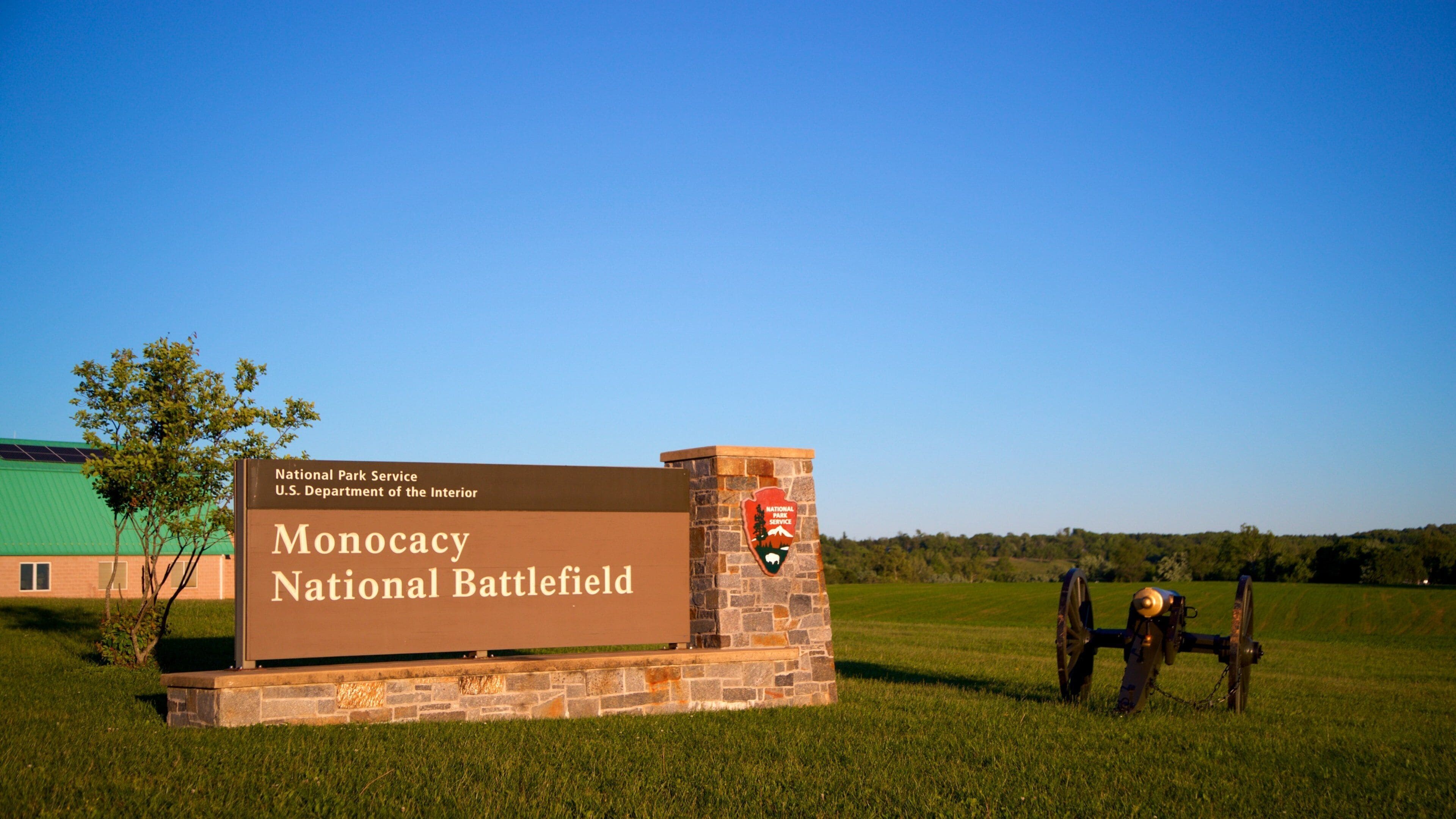 Un cartel del Campo de Batalla Nacional de Monocacy, con una base de piedra y un edificio verde al fondo.