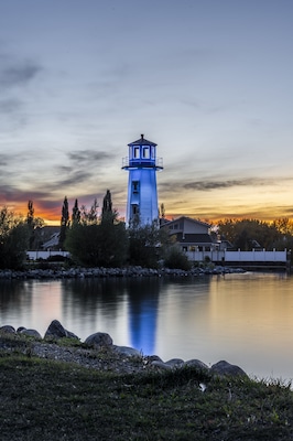 A lighthouse by a calm lake at dusk with a sunset sky.