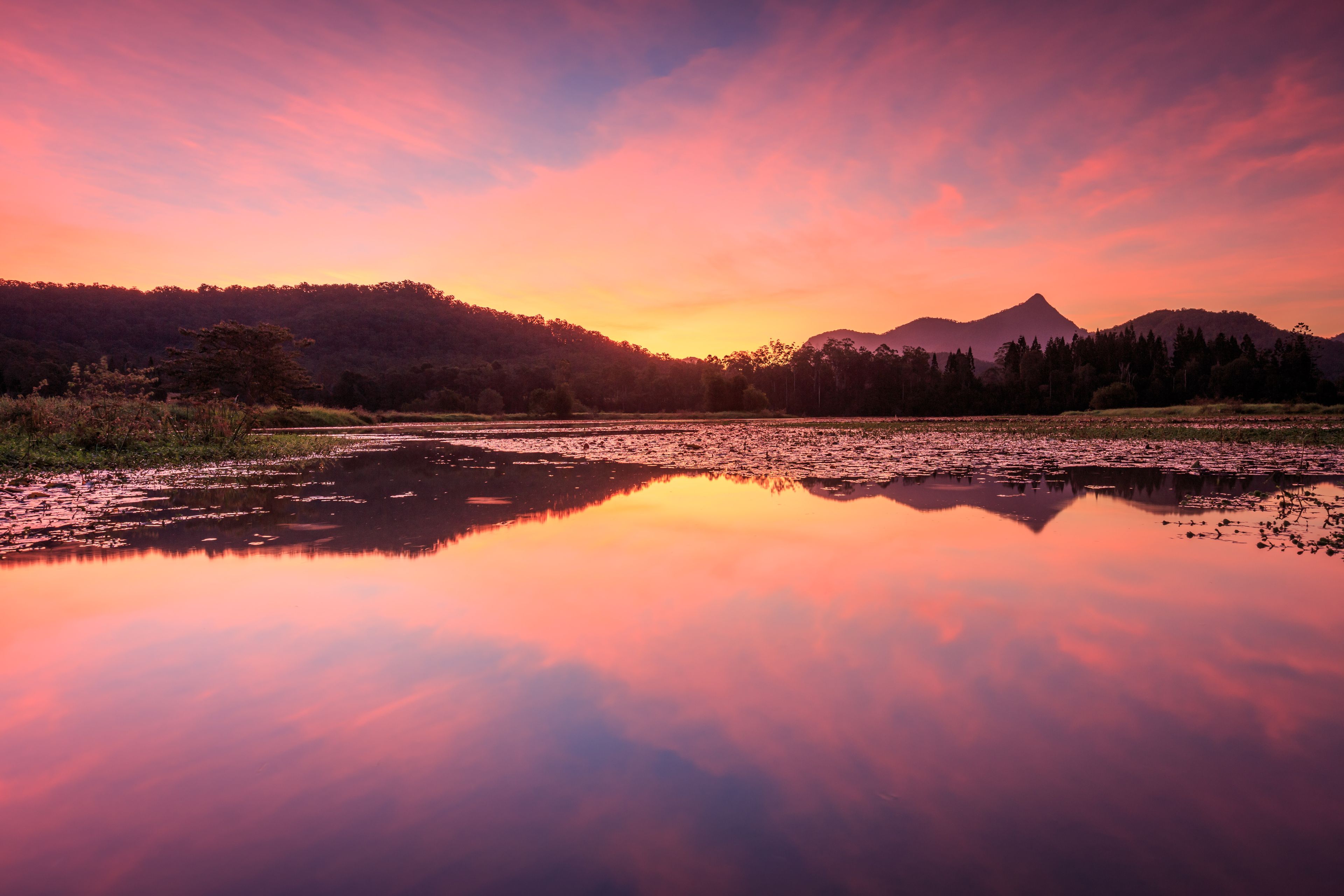 Un lago tranquilo reflejando el cielo al atardecer, rodeado de colinas boscosas y una montaña a lo lejos.