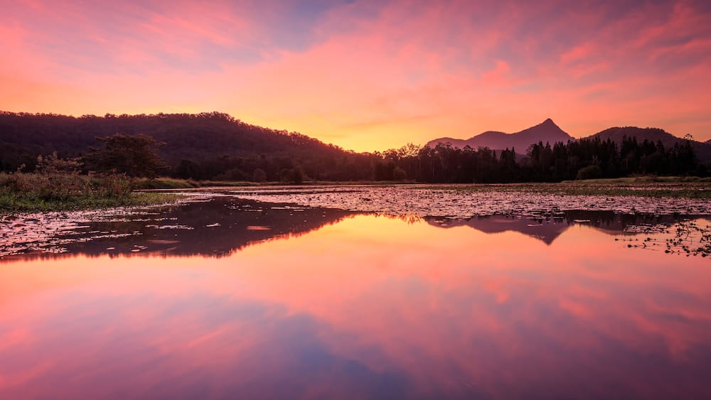 Ein ruhiger See spiegelt den Abendhimmel wider, umgeben von bewaldeten Hügeln und einem Berg in der Ferne.
