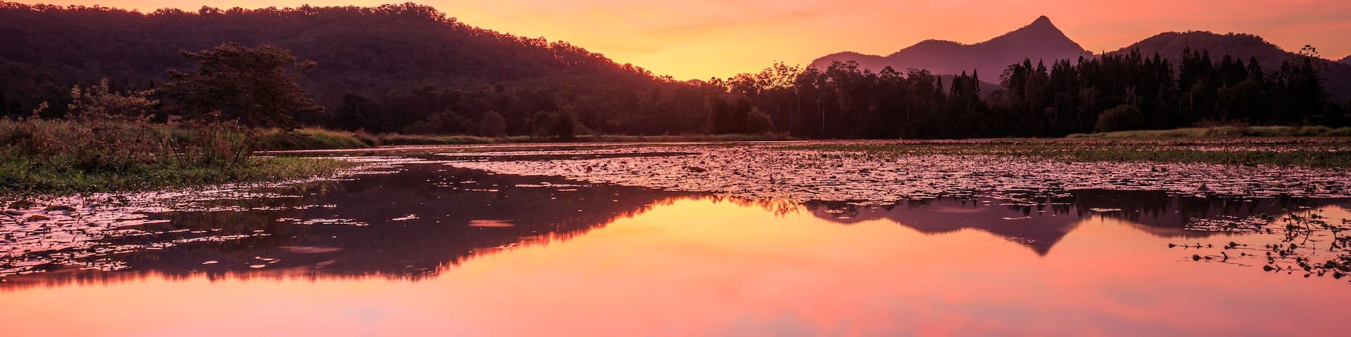 A calm lake reflecting the sunset sky, surrounded by forested hills and a mountain in the distance.