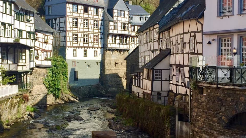 A river flowing through a town with traditional half-timbered houses and a castle in the background.