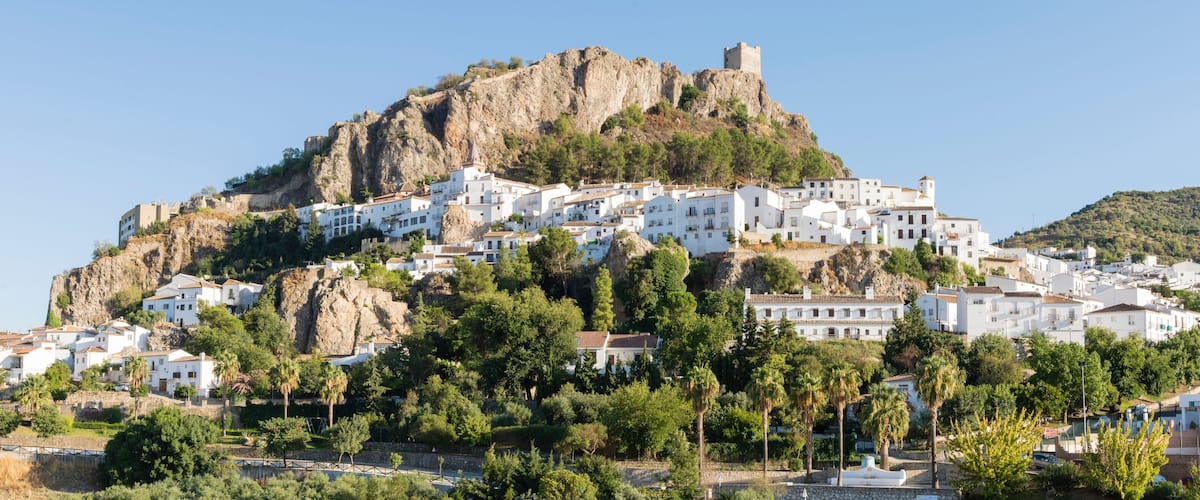 A coastal town with white buildings on a hillside, a prominent rocky peak, and a castle-like structure on top.