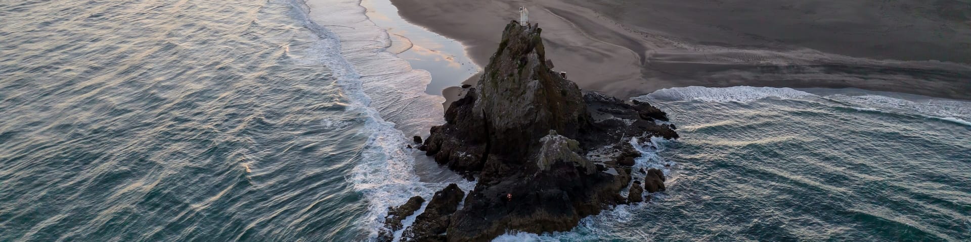 A coastal landscape with a rocky outcrop in the foreground and a sandy beach extending to the ocean.