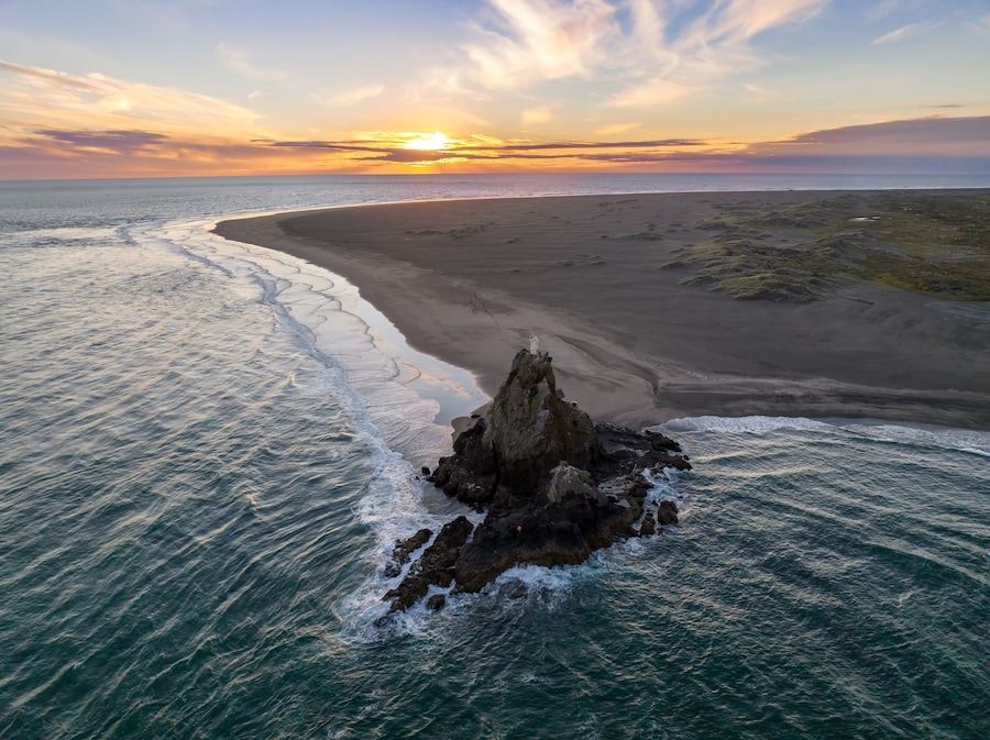 Paisagem costeira com um afloramento rochoso em primeiro plano e uma praia arenosa que se estende até o oceano.