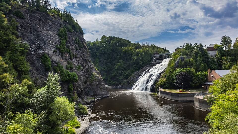 A waterfall cascading down a rocky cliff into a river, surrounded by lush greenery and a bridge.