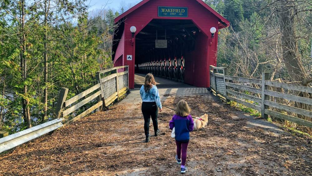 A mother and daughter walking together through the red Wakefield covered bridge along the Gatineau River on a beautiful sunny fall day in pretty town of Wakefield, Quebec, Canada.