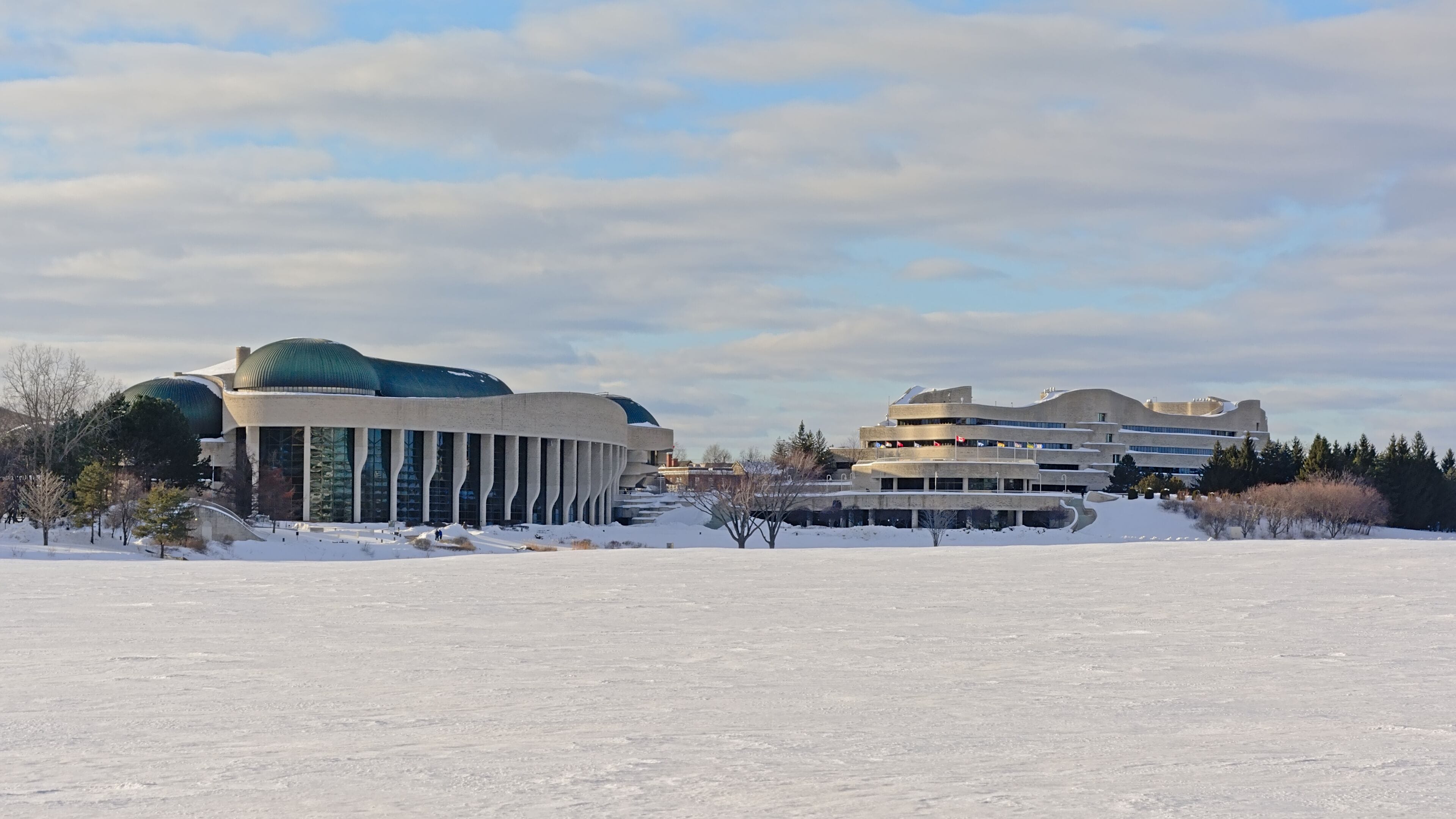 A snow-covered landscape with two modern buildings, one with a dome and another with a curved facade.