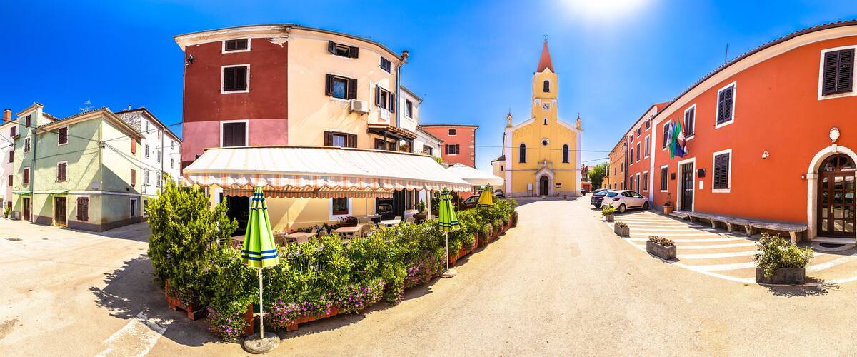 A sunny street with colorful buildings, a church, and outdoor seating with umbrellas.
