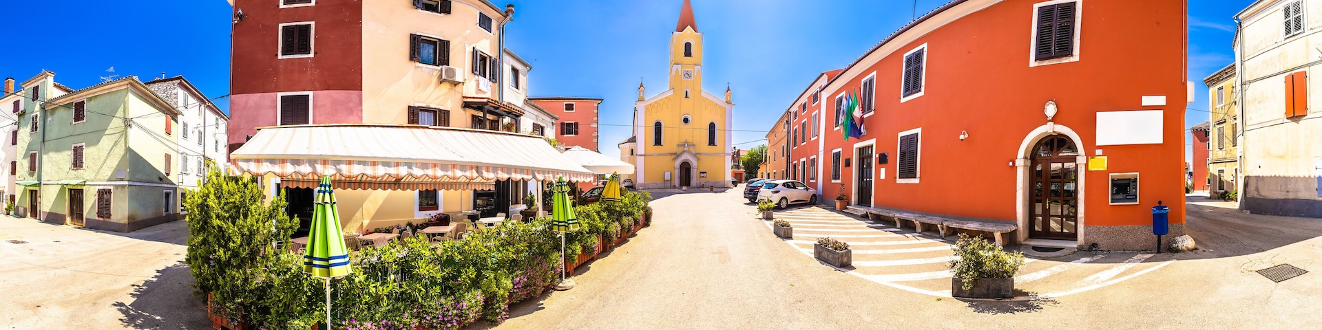 A sunny street with colorful buildings, a church, and outdoor seating with umbrellas.