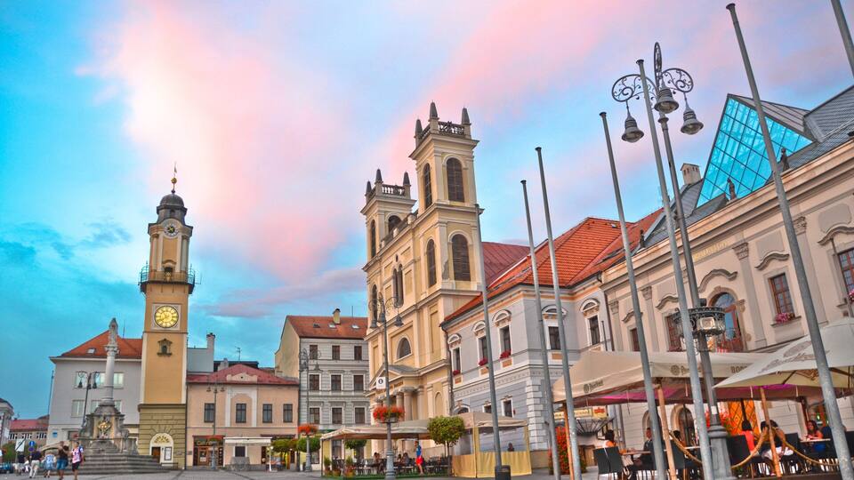 A historic square with a clock tower, a church, and outdoor cafes.