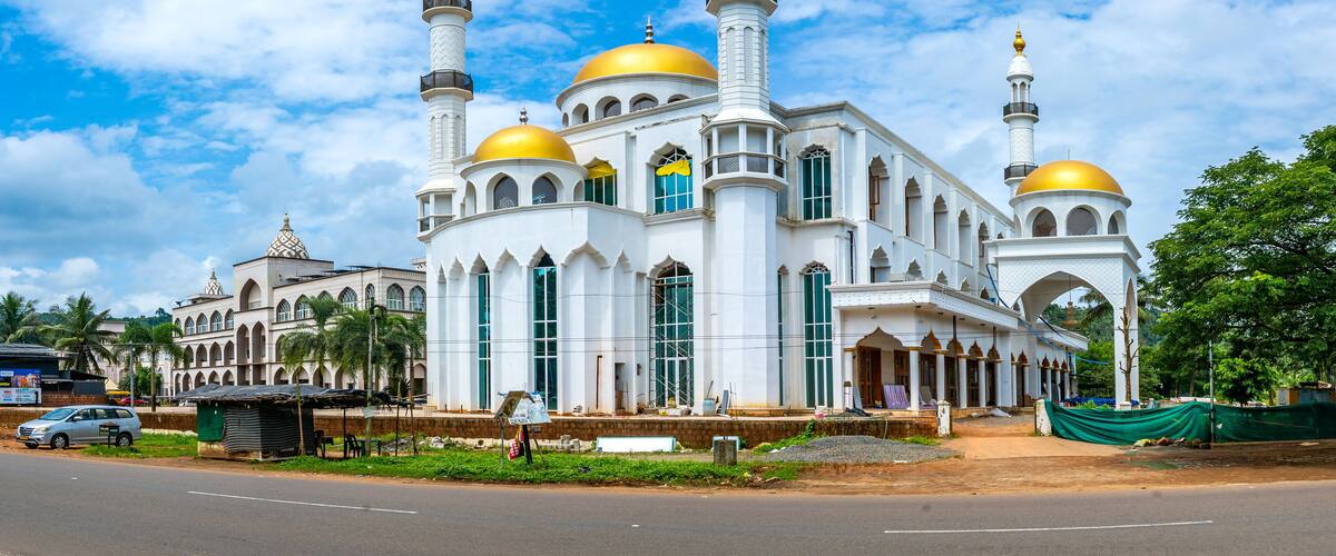 A large white mosque with golden domes and minarets, surrounded by greenery and palm trees.