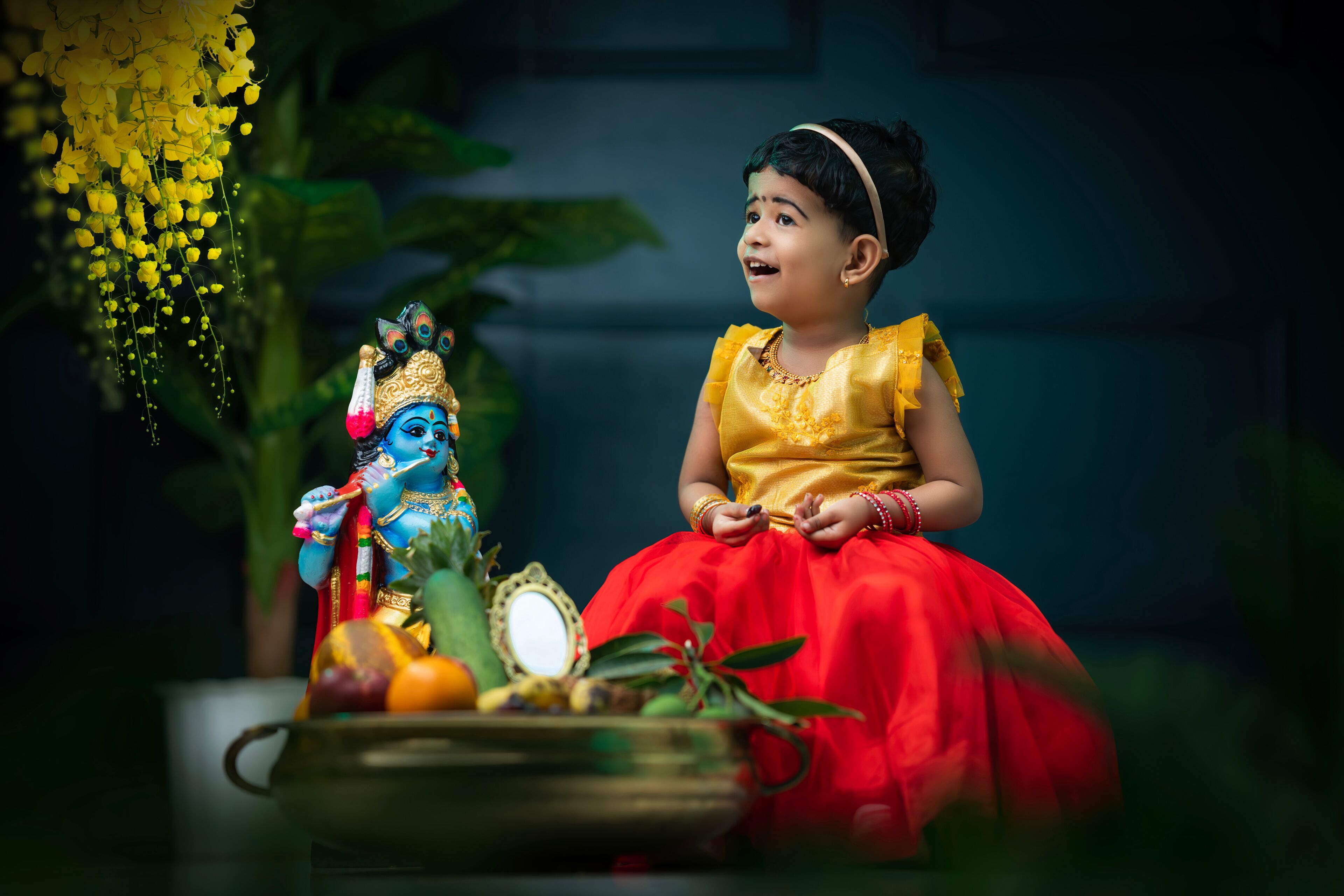 A child in a red dress with a blue deity figure and a mirror on a table.