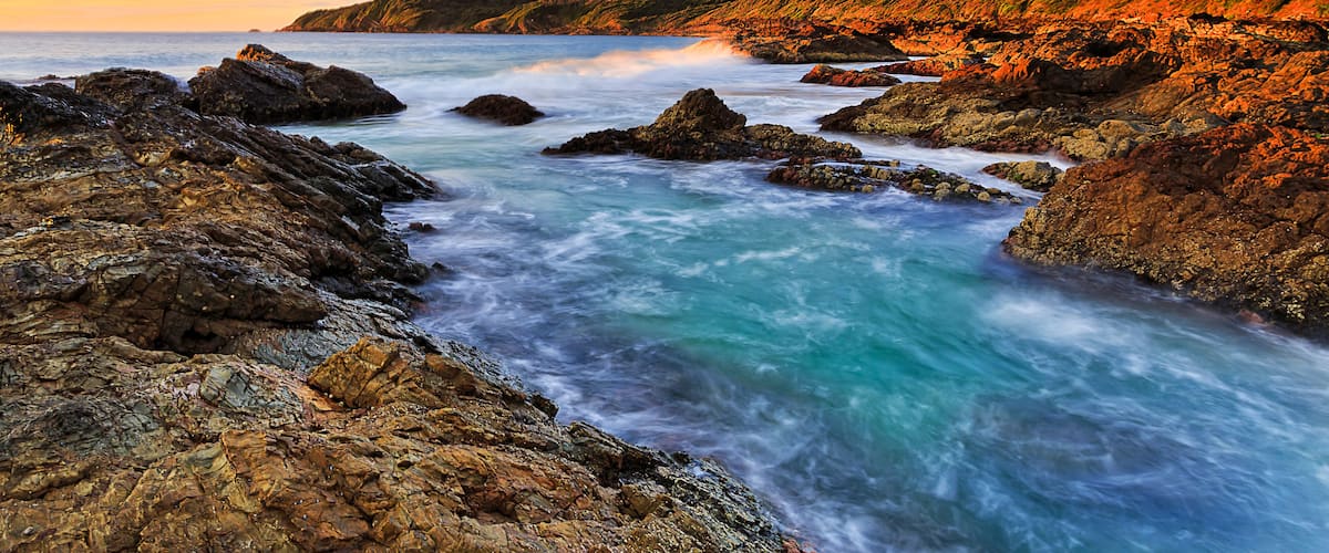 A coastal landscape with rocky shores, clear blue waters, and a hilly backdrop during sunset.