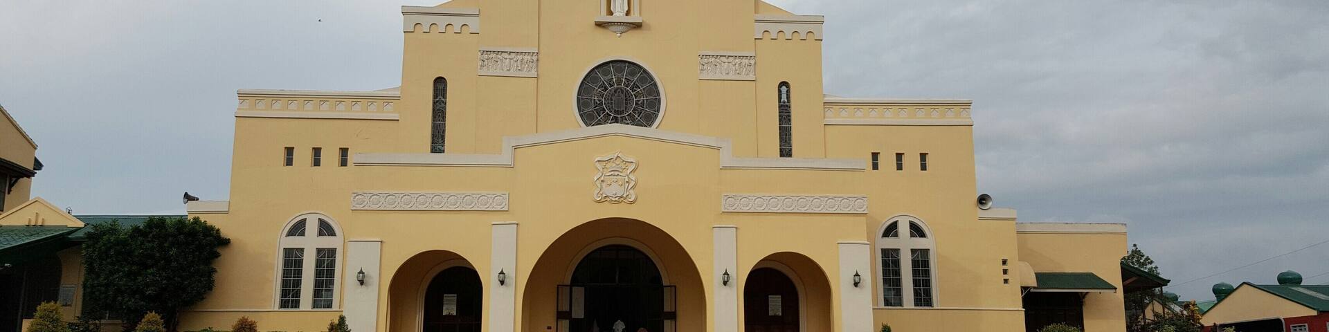 A yellow church with a large circular window, arched entrances, and a cross on top.