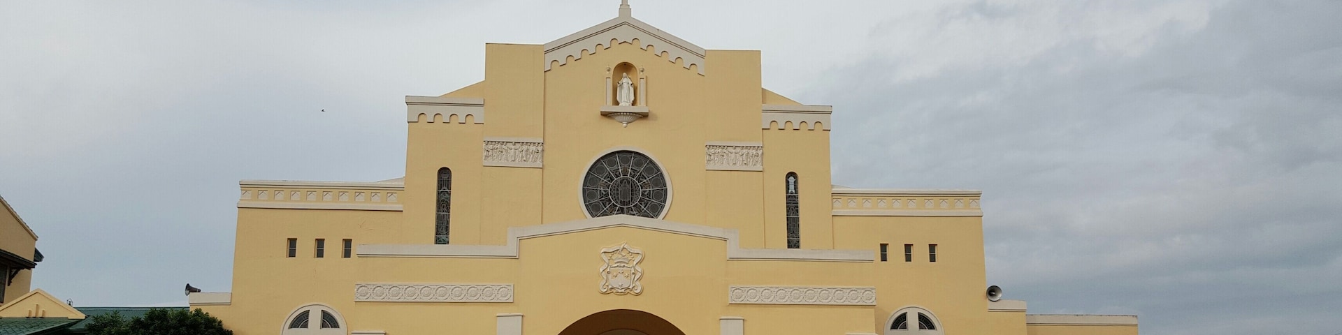 A yellow church with a large circular window, arched entrances, and a cross on top.