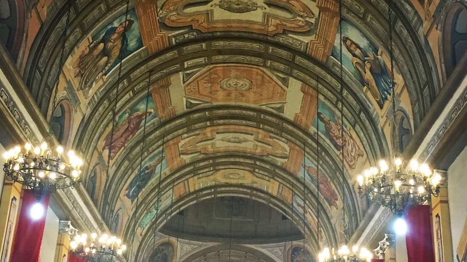 A grand hall with ornate ceiling frescoes, red drapery, and multiple chandeliers.