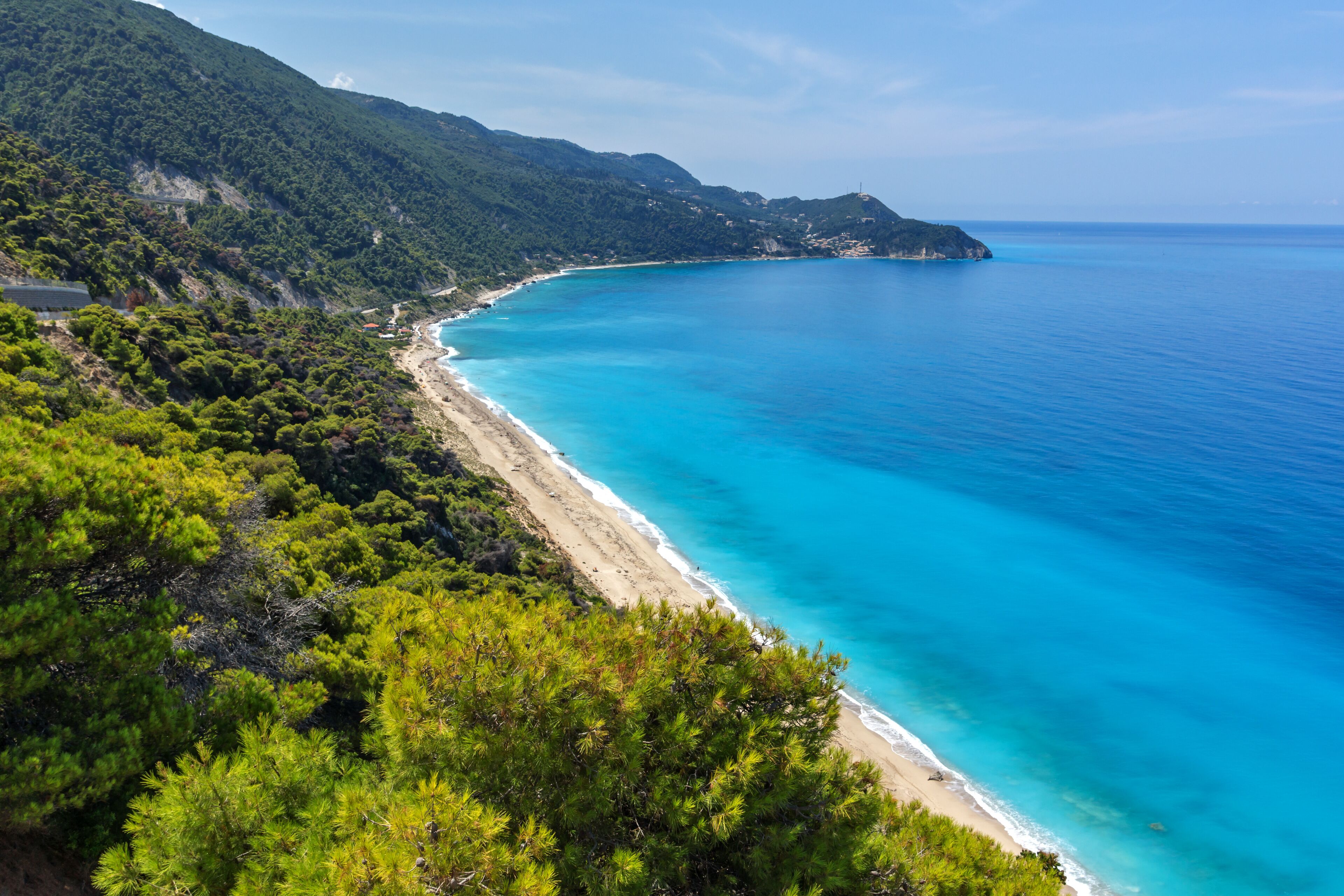 A coastal landscape with a clear turquoise sea, a sandy beach, and dense green vegetation on the hills.