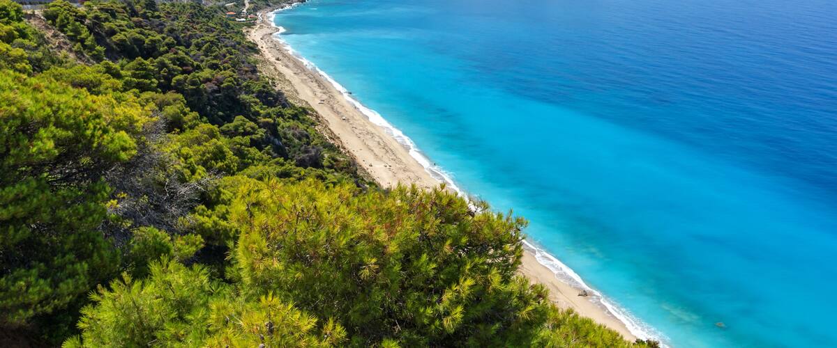 A coastal landscape with a clear turquoise sea, a sandy beach, and dense green vegetation on the hills.